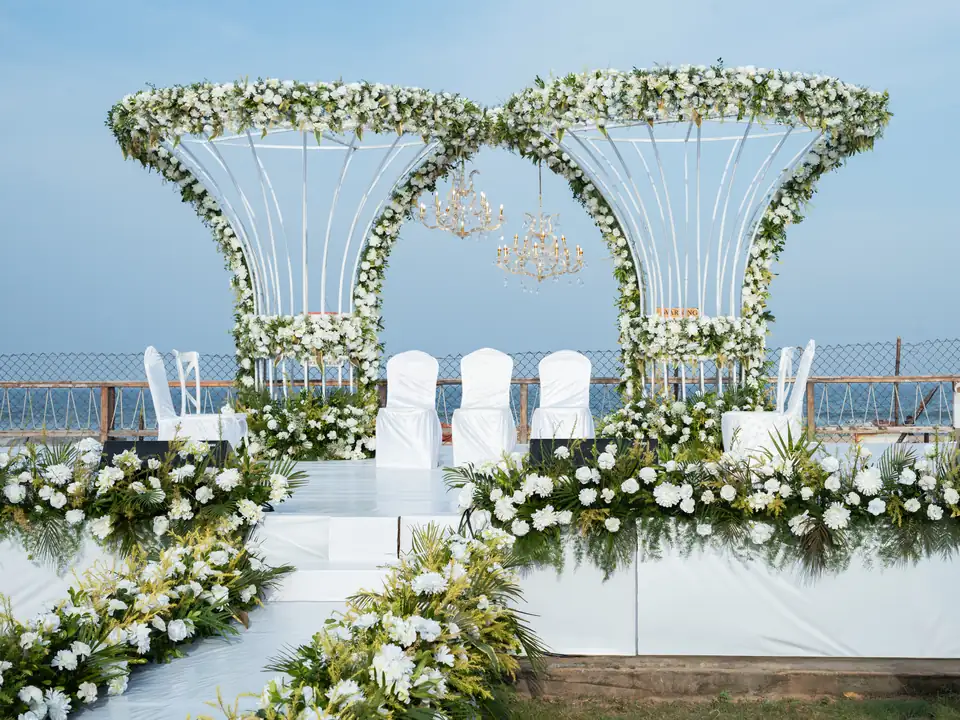 Wedding stage with twin white floral arches, white chairs and greenery, with the sea behind